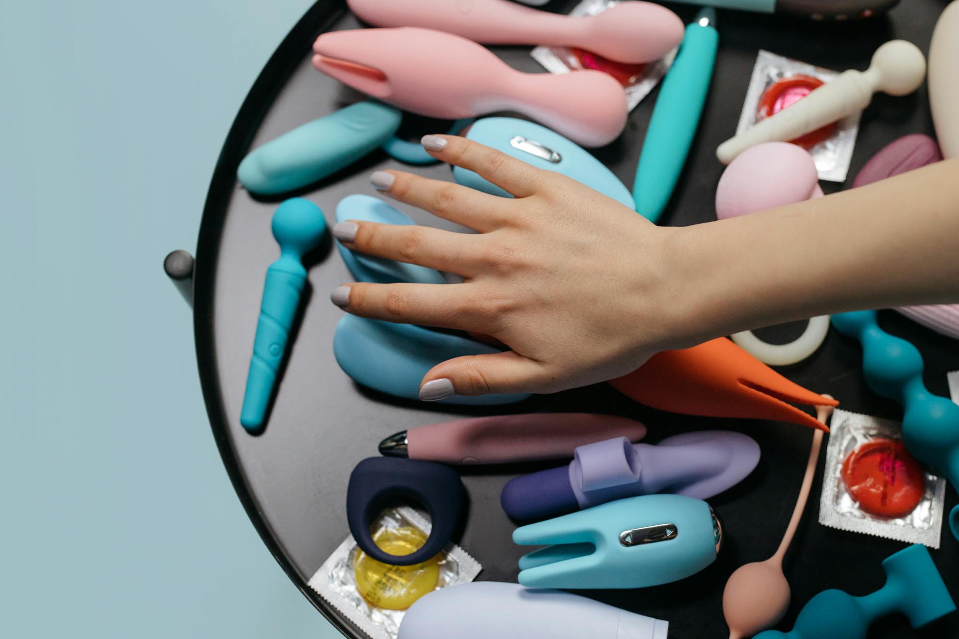 A hand reaching toward a collection of colorful clitoral vibrators arranged on a white surface.
